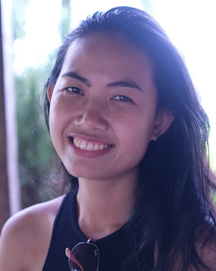 Kunthea smiling at the camera, with long dark hair and a black sleeveless top, outdoors in soft daylight with green leaves behind her.